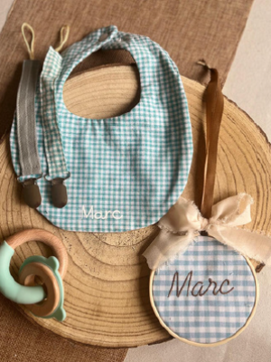 Conjunto de artículos personalizados con el nombre "Marc", que incluye un babero de cuadros celestes, una pinza de chupete, un sonajero de silicona y una decoración circular de tela con lazo y el nombre bordado.