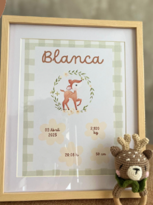 Lámina enmarcada con el nombre "Blanca" en la parte superior, un dibujo de un cervatillo en el centro y detalles del nacimiento como fecha, hora, peso y altura. En primer plano hay un sonajero tejido con forma de reno.