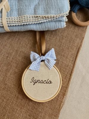 Decoración circular de tela beige con borde de aro de madera, un lazo de cuadros celestes y el nombre "Ignacio" bordado en hilo marrón, acompañado de una manta azul con encaje y un mordedor de madera con tejido azul.